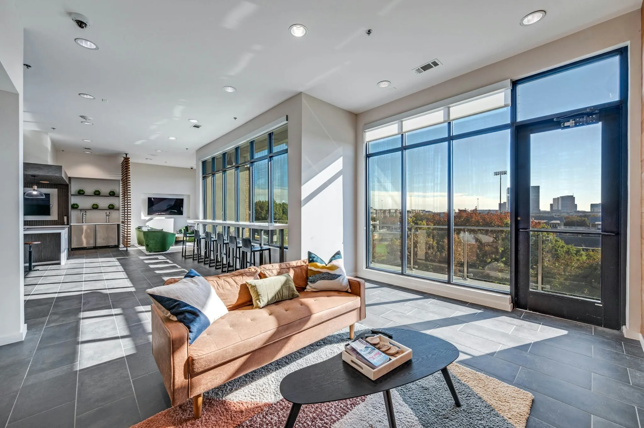 AMLI Frisco Crossing - Spacious lounge area with large windows, bar seating, plush tan sofa over a decorative rug at AMLI Frisco Crossing apartment sky lounge